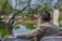 A person in a beige suit is seated outdoors, overlooking a serene pond surrounded by lush greenery and trees. The scene includes an open doorway structure near the water's edge, with a lamp post visible in the background. The person's arm rests on the back of a white chair, creating a relaxed atmosphere.