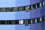 The image depicts a modern building facade with a series of windows. The exterior features a gradient of blue tones, ranging from light blue to dark blue. The windows are framed in black and are uniformly aligned, except for one window with colorful decorations inside.