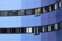 The image depicts a modern building facade with a series of windows. The exterior features a gradient of blue tones, ranging from light blue to dark blue. The windows are framed in black and are uniformly aligned, except for one window with colorful decorations inside.