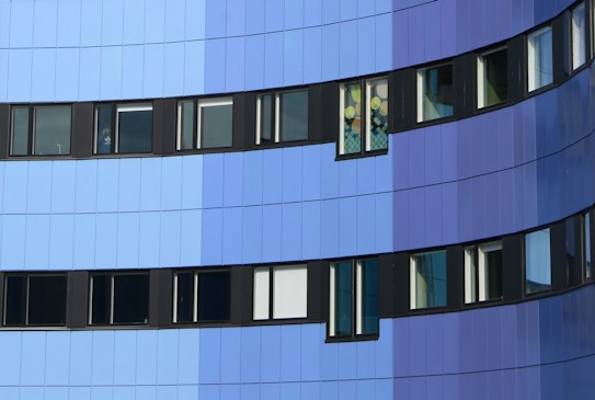 The image depicts a modern building facade with a series of windows. The exterior features a gradient of blue tones, ranging from light blue to dark blue. The windows are framed in black and are uniformly aligned, except for one window with colorful decorations inside.