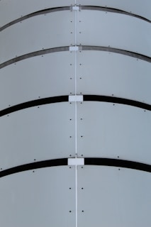 A close-up view of a curved metal surface featuring horizontal bands and lines of rivets. The design includes symmetrical patterns and a minimalist industrial aesthetic.