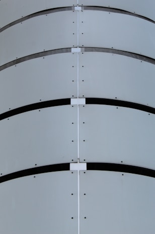 A close-up view of a curved metal surface featuring horizontal bands and lines of rivets. The design includes symmetrical patterns and a minimalist industrial aesthetic.