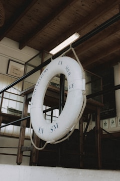 A lifebuoy is hanging from a rope in an indoor setting with wooden beams and glass panels. The text 'SAUVETEURS EN MER GF SN SM' is visible on the lifebuoy. The background includes framed pictures and dark furniture.
