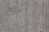 A textured concrete surface with small holes and dents. The surface appears rough and uneven, consisting of various shades of gray with some subtle lines and variations in color and texture.