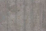 A textured concrete surface with small holes and dents. The surface appears rough and uneven, consisting of various shades of gray with some subtle lines and variations in color and texture.