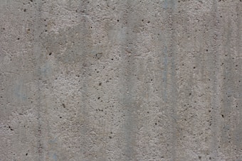 A textured concrete surface with small holes and dents. The surface appears rough and uneven, consisting of various shades of gray with some subtle lines and variations in color and texture.