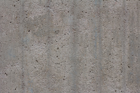 A textured concrete surface with small holes and dents. The surface appears rough and uneven, consisting of various shades of gray with some subtle lines and variations in color and texture.