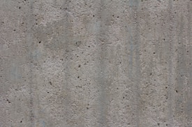 A textured concrete surface with small holes and dents. The surface appears rough and uneven, consisting of various shades of gray with some subtle lines and variations in color and texture.