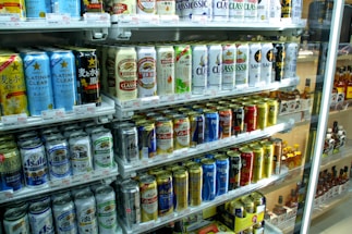 A refrigerated display stocked with a variety of canned beverages, predominantly beer. The cans display colorful designs and different brands, with signs and price tags visible. The shelves are neatly arranged, showcasing a wide selection of drinks for sale.