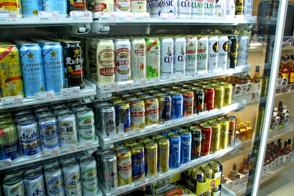 A refrigerated display stocked with a variety of canned beverages, predominantly beer. The cans display colorful designs and different brands, with signs and price tags visible. The shelves are neatly arranged, showcasing a wide selection of drinks for sale.