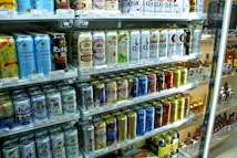 A refrigerated display stocked with a variety of canned beverages, predominantly beer. The cans display colorful designs and different brands, with signs and price tags visible. The shelves are neatly arranged, showcasing a wide selection of drinks for sale.