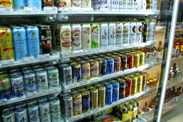 A refrigerated display stocked with a variety of canned beverages, predominantly beer. The cans display colorful designs and different brands, with signs and price tags visible. The shelves are neatly arranged, showcasing a wide selection of drinks for sale.