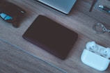 A neatly arranged collection of everyday items on a wooden surface, including a pair of sunglasses, a closed black wallet, white wireless earbuds in their case, and a silver keyring. The corner of a laptop and a pen are partially visible in the background, suggesting a casual or work-related setting.