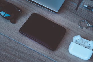 A neatly arranged collection of everyday items on a wooden surface, including a pair of sunglasses, a closed black wallet, white wireless earbuds in their case, and a silver keyring. The corner of a laptop and a pen are partially visible in the background, suggesting a casual or work-related setting.