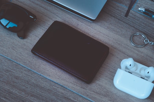 A neatly arranged collection of everyday items on a wooden surface, including a pair of sunglasses, a closed black wallet, white wireless earbuds in their case, and a silver keyring. The corner of a laptop and a pen are partially visible in the background, suggesting a casual or work-related setting.