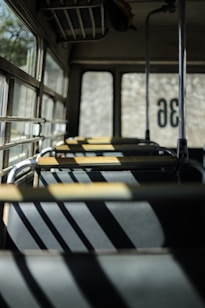 An empty bus interior with sunlight streaming through the windows, casting shadows on the seats. The image focuses on the rows of seats aligned neatly with a vintage metal frame design. Part of the bus window reveals a blurred view of the outside.