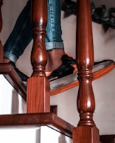 A person is standing on a wooden stairway, wearing rolled-up blue jeans and black sneakers with orange details. The image captures the lower part of the legs and feet. The wood of the stairs is polished, with a rich brown color and intricate grain patterns.