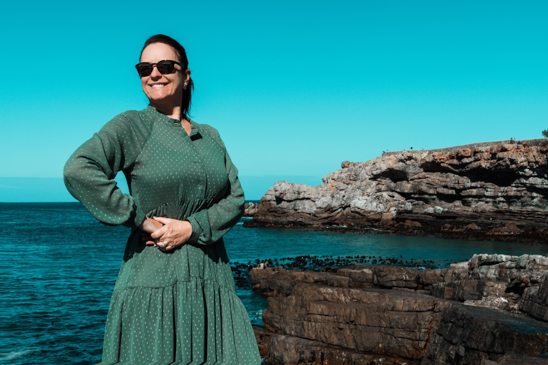 woman in green long sleeve dress wearing black sunglasses standing on rock formation near body of during