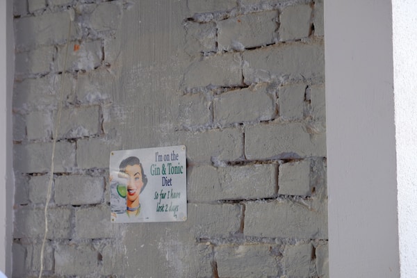 A humorous vintage-style sign is attached to a rough brick wall with faded paint. The sign features a retro illustration of a person smiling while holding a glass, accompanied by a witty caption about a 'Gin & Tonic Diet.'