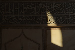Detailed Arabic calligraphy is inscribed on a wall, with sunlight casting a shadow over part of the inscription. The wall is intricately adorned with geometric and floral patterns, demonstrating an artistic and cultural significance.