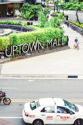 A busy city scene featuring a prominent 'Uptown Mall' sign surrounded by lush greenery. A white taxi is on the road with some signage visible on its side. Several bicycles are parked in the background, and a person is riding a bike on the pathway.
