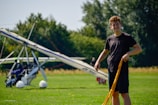 A smiling teen wearing aviation goggles, standing next to a small aircraft on a sunny airfield.
