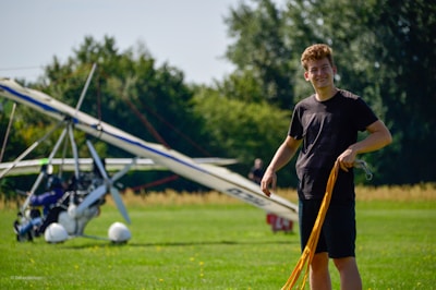 A smiling young man stands on a grassy field, holding a yellow harness or rope. Behind him, a small aircraft with a propeller is parked. The backdrop consists of trees and a clear sky.