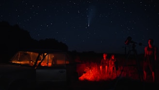 A night sky full of stars with a visible comet. Two people are sitting on the ground near a red-lit car, while another stands beside a telescope. The scene is set in an open field with dark silhouettes of trees in the background.
