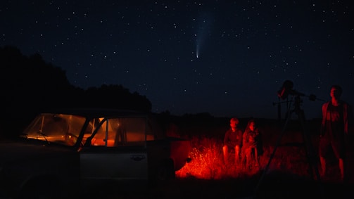 A night sky full of stars with a visible comet. Two people are sitting on the ground near a red-lit car, while another stands beside a telescope. The scene is set in an open field with dark silhouettes of trees in the background.