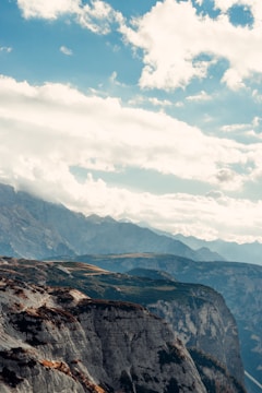 A stunning landscape featuring a dramatic cliff edge with layered rock formations. In the background, a series of majestic mountains stretches across the horizon under a partly cloudy sky.