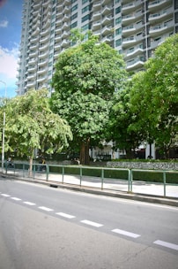 A tall, modern high-rise apartment building with numerous balconies stands in the background. In the foreground, there is a well-maintained sidewalk lined with lush, green trees. A metal railing separates the sidewalk from a paved road, which features clearly marked white lines.