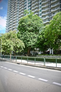 A tall, modern high-rise apartment building with numerous balconies stands in the background. In the foreground, there is a well-maintained sidewalk lined with lush, green trees. A metal railing separates the sidewalk from a paved road, which features clearly marked white lines.