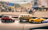 A collection of toy model cars sits on the dashboard of a vehicle, featuring a variety of designs including a red Mini Cooper, a beige Volkswagen camper van, and a yellow sports car. The background is blurred, suggesting an outdoor urban setting, possibly a street with buildings.