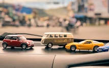 A collection of toy model cars sits on the dashboard of a vehicle, featuring a variety of designs including a red Mini Cooper, a beige Volkswagen camper van, and a yellow sports car. The background is blurred, suggesting an outdoor urban setting, possibly a street with buildings.