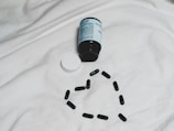 A black bottle of pills is lying on its side on a white surface, with its white cap detached. Several black capsules are arranged in the shape of a heart below the bottle.