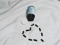 A black bottle of pills is lying on its side on a white surface, with its white cap detached. Several black capsules are arranged in the shape of a heart below the bottle.