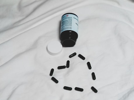 A black bottle of pills is lying on its side on a white surface, with its white cap detached. Several black capsules are arranged in the shape of a heart below the bottle.