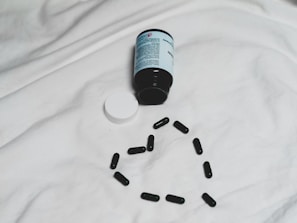A black bottle of pills is lying on its side on a white surface, with its white cap detached. Several black capsules are arranged in the shape of a heart below the bottle.