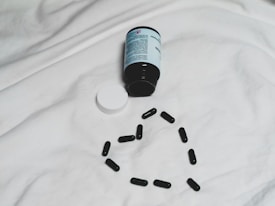 A black bottle of pills is lying on its side on a white surface, with its white cap detached. Several black capsules are arranged in the shape of a heart below the bottle.