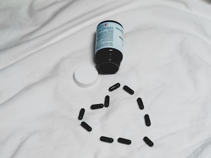 A black bottle of pills is lying on its side on a white surface, with its white cap detached. Several black capsules are arranged in the shape of a heart below the bottle.