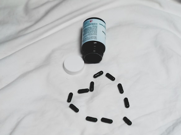 A black bottle of pills is lying on its side on a white surface, with its white cap detached. Several black capsules are arranged in the shape of a heart below the bottle.