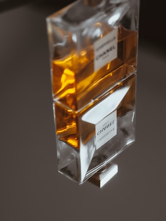 A transparent perfume bottle containing an amber-colored liquid is displayed elegantly on a reflective surface. The label on the bottle is visible through the glass.