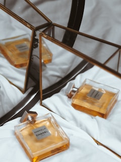 A bottle of Chanel perfume is placed on a soft, white fabric surface. Its elegant design features a square glass bottle with an amber-colored fragrance, creating a luxurious and sophisticated impression. The perfume bottle is reflected in a geometric, triangular mirror, adding depth and complexity to the composition.