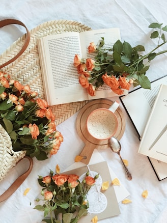 An arrangement featuring an open book surrounded by peach-colored roses, lying on a woven basket. A cup of frothy coffee sits on a wooden coaster beside a spoon. Scattered yellow petals and a decorative card add to the cozy, inviting atmosphere.