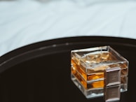 A square glass perfume bottle with an amber-colored liquid placed on a reflective black surface, accompanied by a small, square object with a logo.