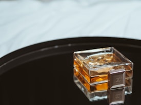A square glass perfume bottle with an amber-colored liquid placed on a reflective black surface, accompanied by a small, square object with a logo.