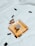 A bottle of Chanel perfume lies on a soft, white fabric. Several black capsules are scattered around it, adding a contrast to the clean and minimalist background. The perfume bottle is square-shaped with a golden hue, reflecting elegance and luxury.