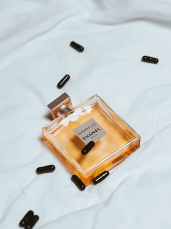 A bottle of Chanel perfume lies on a soft, white fabric. Several black capsules are scattered around it, adding a contrast to the clean and minimalist background. The perfume bottle is square-shaped with a golden hue, reflecting elegance and luxury.