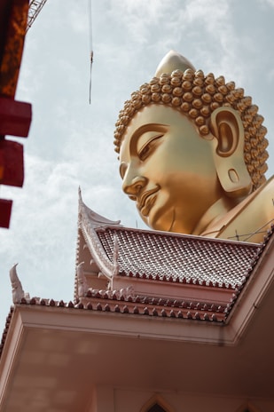 A large golden Buddha statue head overlooks a traditional Thai temple roof. The intricate details of the statue's curls and serene expression stand out against a cloudy sky. The temple roof features ornate carvings and a patterned design.