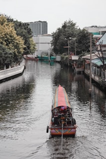 A long-tail boat travels down a narrow canal flanked by trees and buildings. The water reflects the colors of the environment, creating a serene setting. The background features urban structures, including a tall building.